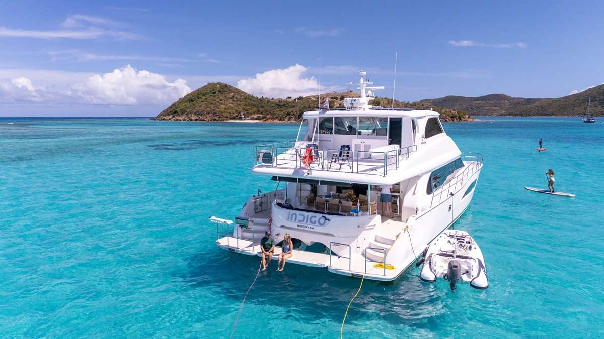 Indigo at anchor in a turquoise BVI bay, guests on the swim platform and paddleboards in the water
